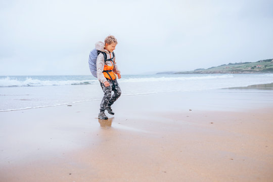 Boy little backpacker runs from ocean waves on the deserted beach in rainy day