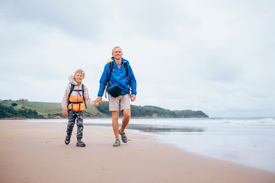 Traveling with child: father and son walk with backpack on sandy ocean beach, rainy day