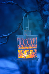 Decorative Christmas lantern with burning candle hanging on snow-covered fir- tree branch in a winter park. New year festive card, poster, postcard design.