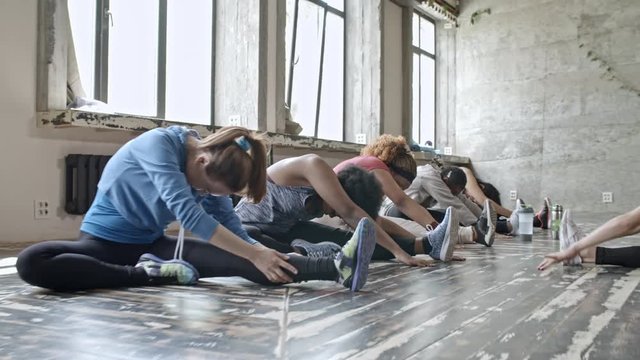 PAN of curly female instructor and group of young women stretching legs and bending over while sitting on floor in studio during flexibility class