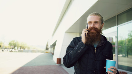 happy beard young man talking on the smartphone and drinking coffee while walking in the street