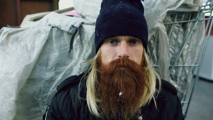 Closeup portrait of young bearded homeless man sitting on a sidewalk near shopping cart ang garbage container during cold winter day