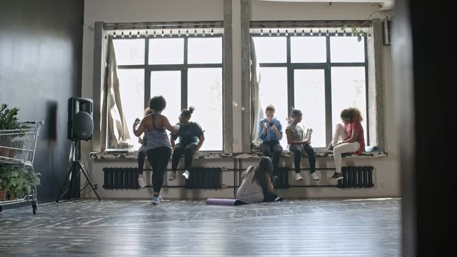 Wide shot of multiethnic group of women sitting on windowsills and chatting before fitness class