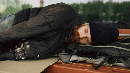 Young Homeless man trying to sleep under jacket on bench at the sidewalk