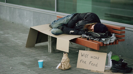 Young homeless drunk man preparing to sleep on cardboard on bench at the sidewalk