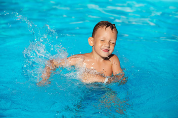 Fototapeta na wymiar Happy kid playing in blue water of swimming pool. Little boy learning to swim. Summer vacations concept. Cute boy swimming in pool water. Child splashing in swimming pool