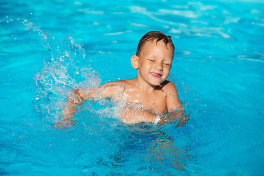 Happy kid playing in blue water of swimming pool. Little boy learning to swim. Summer vacations concept. Cute boy swimming in pool water. Child splashing in swimming pool