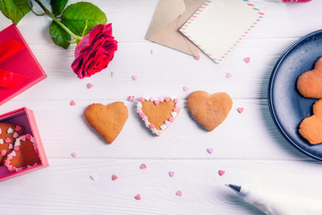 Homemade decorating cookies in shape of heart as gift for lover on Valentine's day. White wooden table with greeting card, rose and decor. Festive gift concept. Selective focus. Copy space