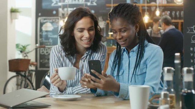 Two Beautiful Girlfriends Sitting in the Coffee Shop and Chatting, One Shows Pictures on Her Smartphone, they Both Laugh. In the Background Stylish Establishment.