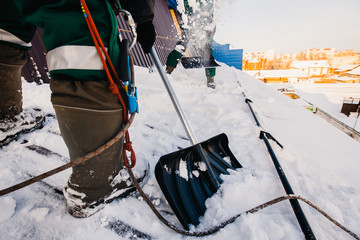 Team of male workers clean roof of building from snow with shovels in securing belts of mantra.