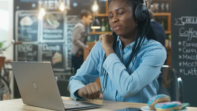 Stylish Young African-American Woman Sits in the Coffee Shop with Her Laptop and Headphones on, Listening to Music and Dancing to the Beats. Shot on RED EPIC-W 8K Helium Cinema Camera.