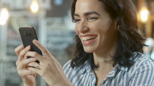 Close-up Portrait Shot of a Beautiful Young Woman Smiling and Using Mobile Phone. In the Background Stylish Coffee Shop. Shot on RED EPIC-W 8K Helium Cinema Camera.