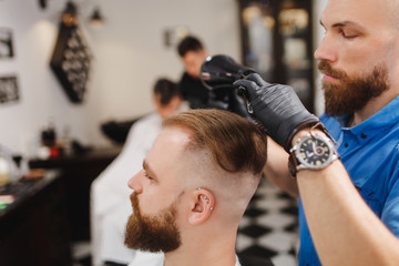 Male professional hairdresser serving client, drying hair with hairdryer. Ginger handsome brutal stylish young man with thick big beard and short hair getting trendy haircut. Light white barber shop.