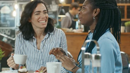Two Beautiful Women Sitting at the Cafe, Drinking Beverages Eating Desserts. They Chat and Have Fun Together. In the Background Modern Stylish Coffee House. Shot on RED EPIC-W 8K Helium Cinema Camera.