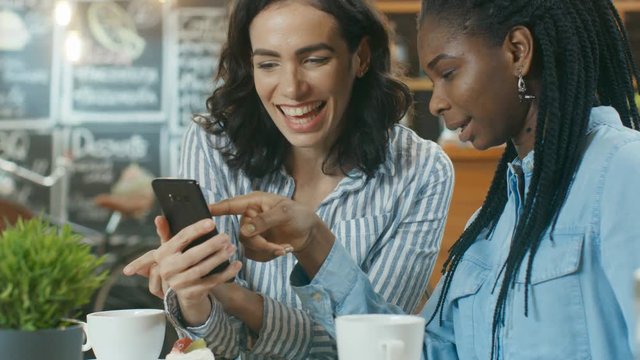 Two Gorgeous Girlfriends Have Fun while Sitting in the Cafe, One Shows Something on a Mobile Phone. In the Background Stylish Modern Cafe. Shot on RED EPIC-W 8K Helium Cinema Camera.