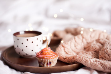 Cupcake with cup of coffee with knitted cloth on wooden tray in bed close up. Good morning.