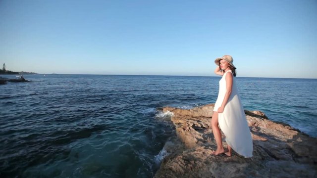 beautiful girl admires the sea. Beautiful girl in a white dress and hat stands on a stone coast near the sea. The concept of an excellent holiday in Greece.