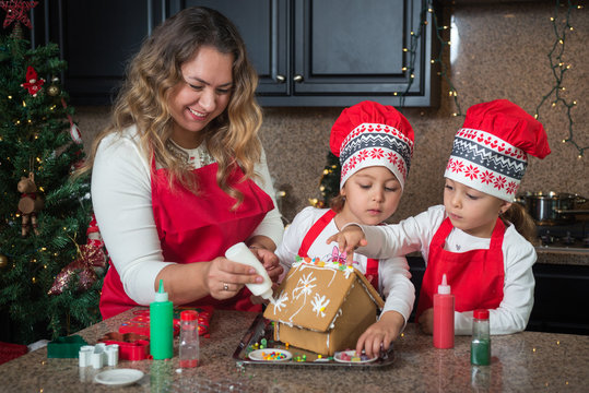 Happy Mom and twin girls in red making Christmas gingerbread house