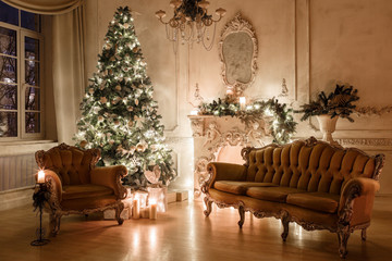 Christmas evening by candlelight. classic apartments with a white fireplace, decorated tree, sofa, large windows and chandelier.