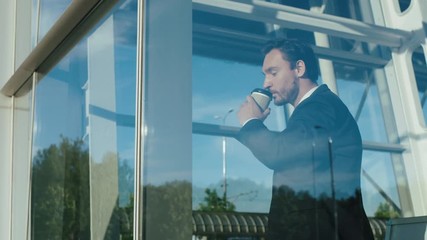 handsome businessman in a tie and suit drinking coffee to go and looking woth a smile out of the window of his office and than going away. Sunny day. Indoor - Powered by Adobe
