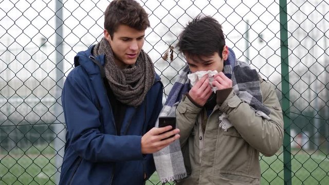Two trendy casual young men standing together outdoors reading an sms or text message, or looking at photos on a mobile phone, surfing the web