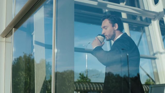 handsome businessman in a tie and suit drinking coffee to go and looking woth a smile out of the window of his office and than going away. Sunny day. Indoor