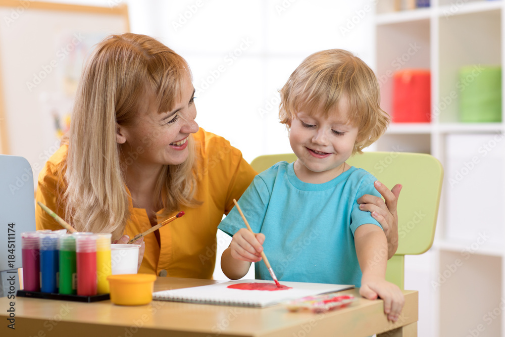 Wall mural Child painting in preschool. Teacher help by little boy.