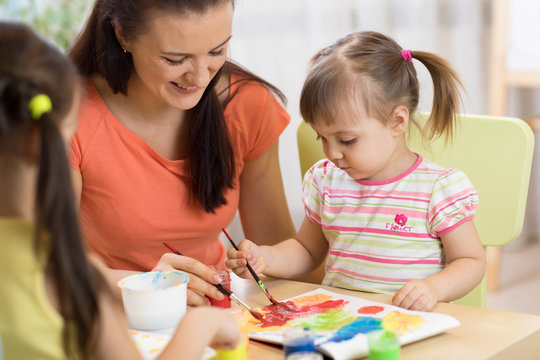 Teacher and little girls are painting in daycare center. Woman and children have a fun pastime.