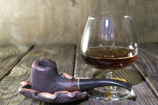 A Smoking pipe in the ashtray and a glass of cognac on oak textured table.