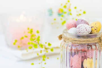 Chocolate Candy Multi-Colored Small Quail Easter Eggs Pastel Colors in Vintage Glass Jar Tied with Twine on White Wood Table Yellow Spring Flowers Lit Candle Copy Space Festive Setting