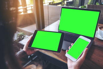 Mockup image of a businesswoman holding white mobile phone , black tablet and laptop with blank green screen on vintage wooden table with coffee cup in cafe - Powered by Adobe