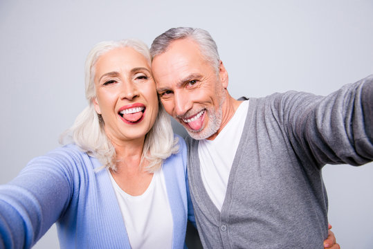 Crazy funky funny elderly couple are taking selfie using smartphone and showing tongues, isolated on grey background