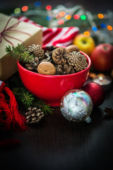 Christmas decoration - red bowl full of fir-cones, gift box wrapped in kraft paper, pine branches, candle, nuts, anise, apples and christmas toysmas= Christmas theme. Dark wooden background.