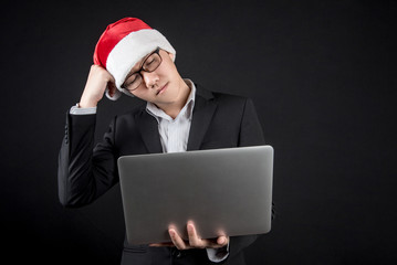 Young Asian businessman wearing suit and santa hat feeling stressed while using laptop computer. male entrepreneur in christmas holiday and new year party concepts on black background