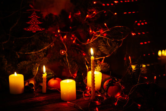 One candle sits in a brass candle holder on a table on Christmas. This short, lit candle is surrounded by green garland and ornaments, in front of blurry lights.