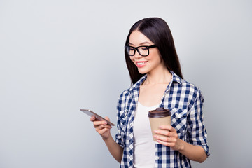 Close up portrait of happy girl in glasses searching something in her telephone through 5g internet in one hand , holding cup in her second one  while standing over grey background