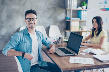 Portrait of handsome bearded attractive young businessman in glasses sitting at his desktop and smiling at camera in open space office - obrazy, fototapety, plakaty