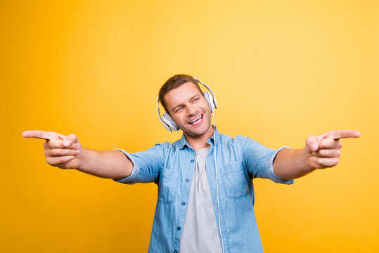 Portrait of caucasion, positive guy in jeans shirt with bristle listening his favorite music, singing the song, pointing index fingers front over yellow background