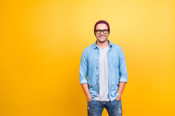 Portrait with copy space of smiling, happy guy in jeans wear, glasses, red cap holding hands in pocket, looking at camera over yellow background