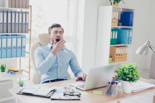 Portrait of sleepy tired agent wearing blue shirt, he is yawning and covering his mouth with a palm, he is sitting in front of computer