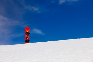 Distant view to red - orange board with black duck stance positioned bindings for snowboarding stuck straight into the snow with background of blue sky 