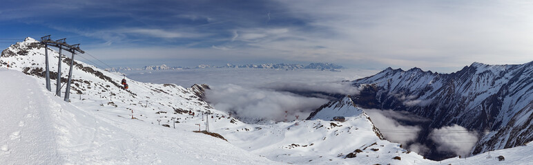Panoramic view of foggy Alps Mountains and cable cars lift with blue sky and white clouds in background at high-altitude ski resort in March