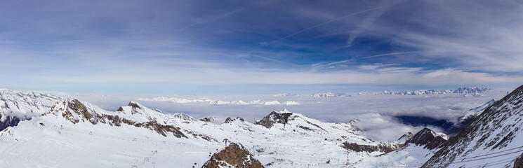 Panorama of high-altitude ski slopes and cable car lifts with foggy Alps mountains, blue sky and white clouds in background at glacier hills in March month