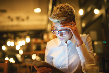 Close up of stylish worried entrepreneur with eyeglasses and earphones looking in a tablet while sitting in a cafe in the night.