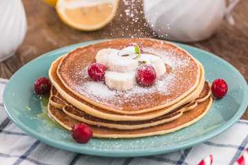 Pancake stack covered with honey, powdered sugar, banana and cranberries. On a wooden background in a blue plate