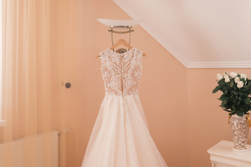 The perfect wedding white lace dress hanging on rack. Beautiful bridal dress on a hanger in the room of the bride over beige wall. Vase of white tender roses. Fashion look