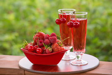 Ripe strawberry, cherry, raspberry in the red bowl, two glasses of soda on a green blurred background. Sunny summer background with different berries and drink red. Shallow depth of field. Selective f