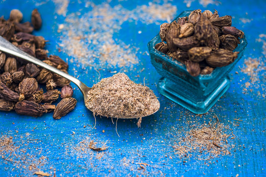 Raw black cardamom on a bluish surface with its powder.;