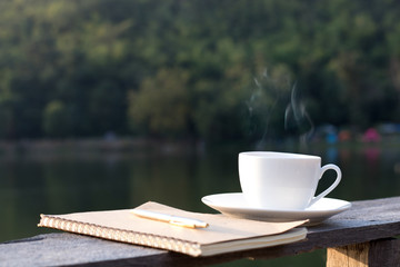 cup of hot coffee for brackfast and note book with pen on wooden balcony near river and mountain background.