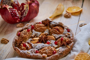 A powdered pear pie decorated with white cloth, fresh sliced pears and pomegranate, dried oranges, walnuts, anise stars on the wooden board. Healthy homemade pear pie with fresh red pomegranate seeds
